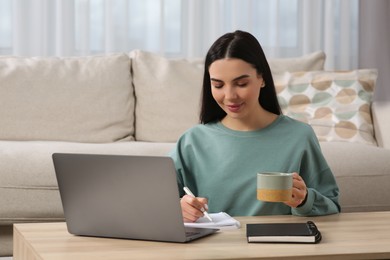 Photo of Happy woman with cup of drink taking notes while working on laptop at wooden desk in living room