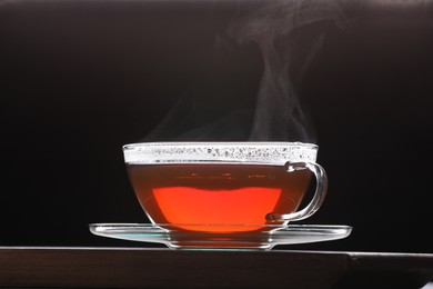 Glass cup of tea with saucer on wooden table against black background