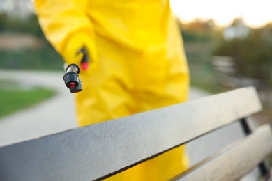 Photo of Person in hazmat suit disinfecting bench in park with sprayer, closeup. Surface treatment during coronavirus pandemic