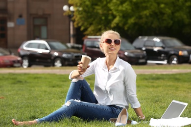 Young woman with cup of coffee sitting on green lawn in park. Joy in moment