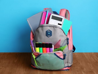 Photo of Backpack with different school stationery on wooden floor near light blue wall