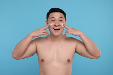 Happy man applying cream onto his face on light blue background