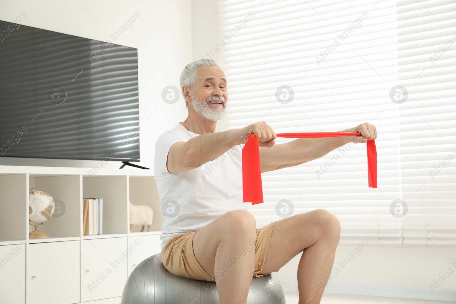 Photo of Senior man doing exercise with elastic resistance band on fitness ball at home