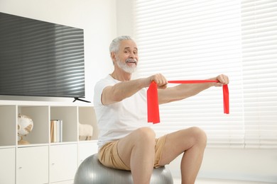 Senior man doing exercise with elastic resistance band on fitness ball at home