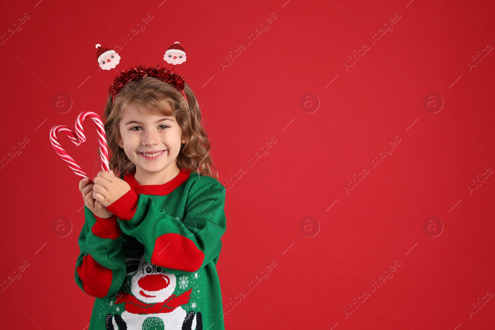 Photo of Cute little girl in Christmas sweater making heart shape with candy canes against red background. Space for text