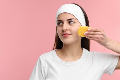 Young woman with headband washing her face using sponge on pink background, space for text