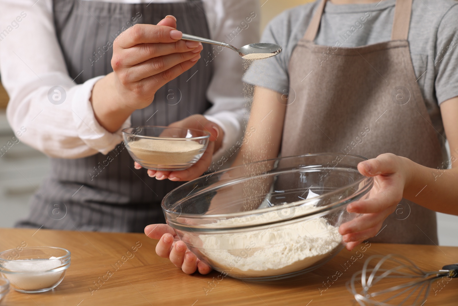 Photo of Making bread. Mother and her daughter putting flour and dry yeast into bowl at wooden table in kitchen, closeup