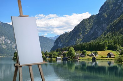 Image of Wooden easel with blank canvas on table and beautiful mountain landscape. Space for text