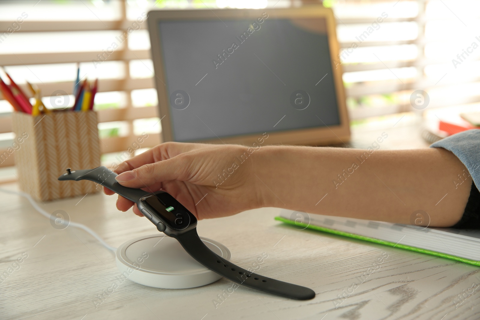 Photo of Woman putting smartwatch onto wireless charger at white wooden table, closeup. Modern workplace accessory