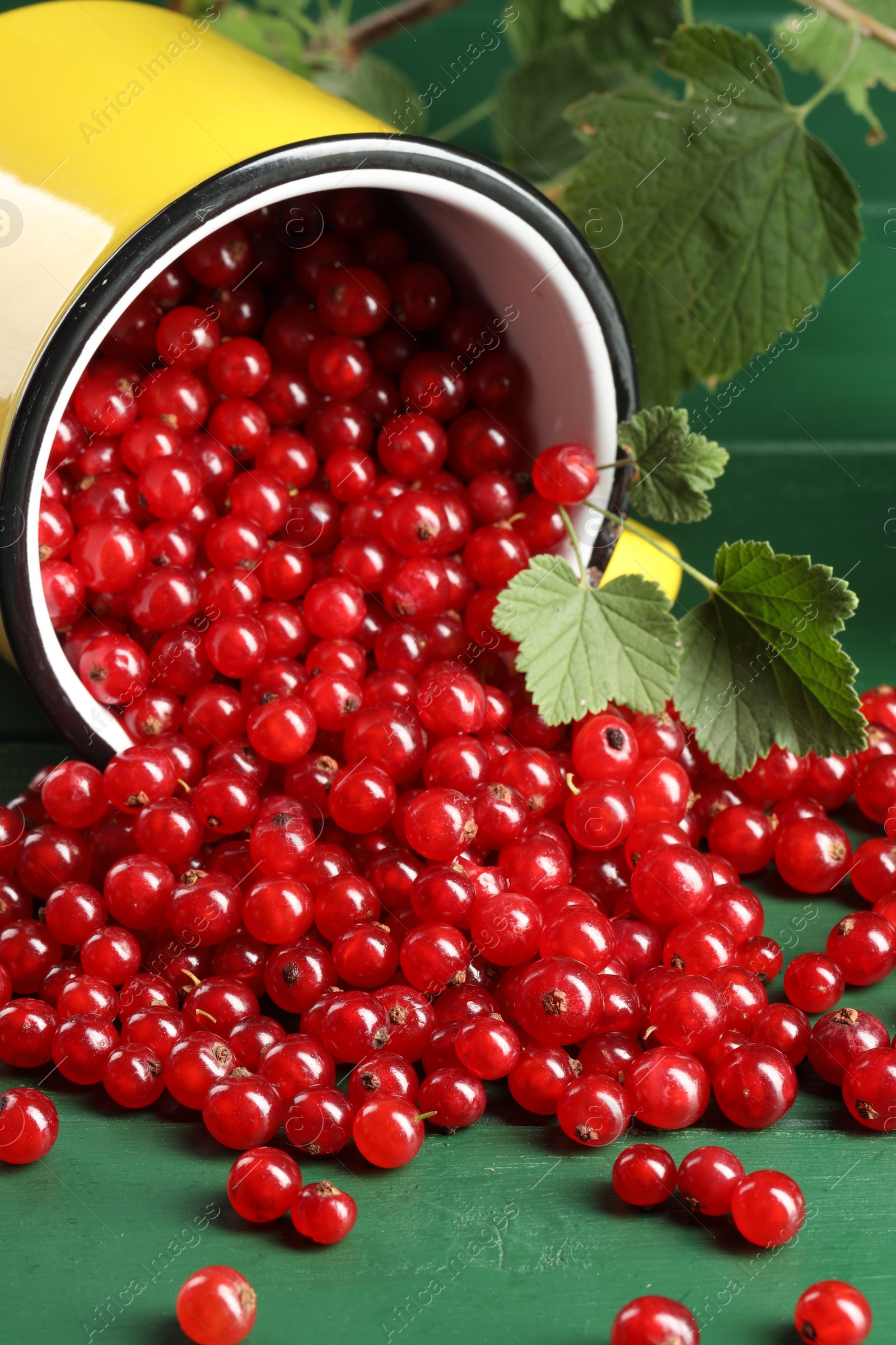 Photo of Many ripe red currants, mug and leaves on green wooden table, closeup
