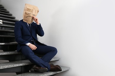 Photo of Man wearing paper bag with drawn sad face indoors. Space for text
