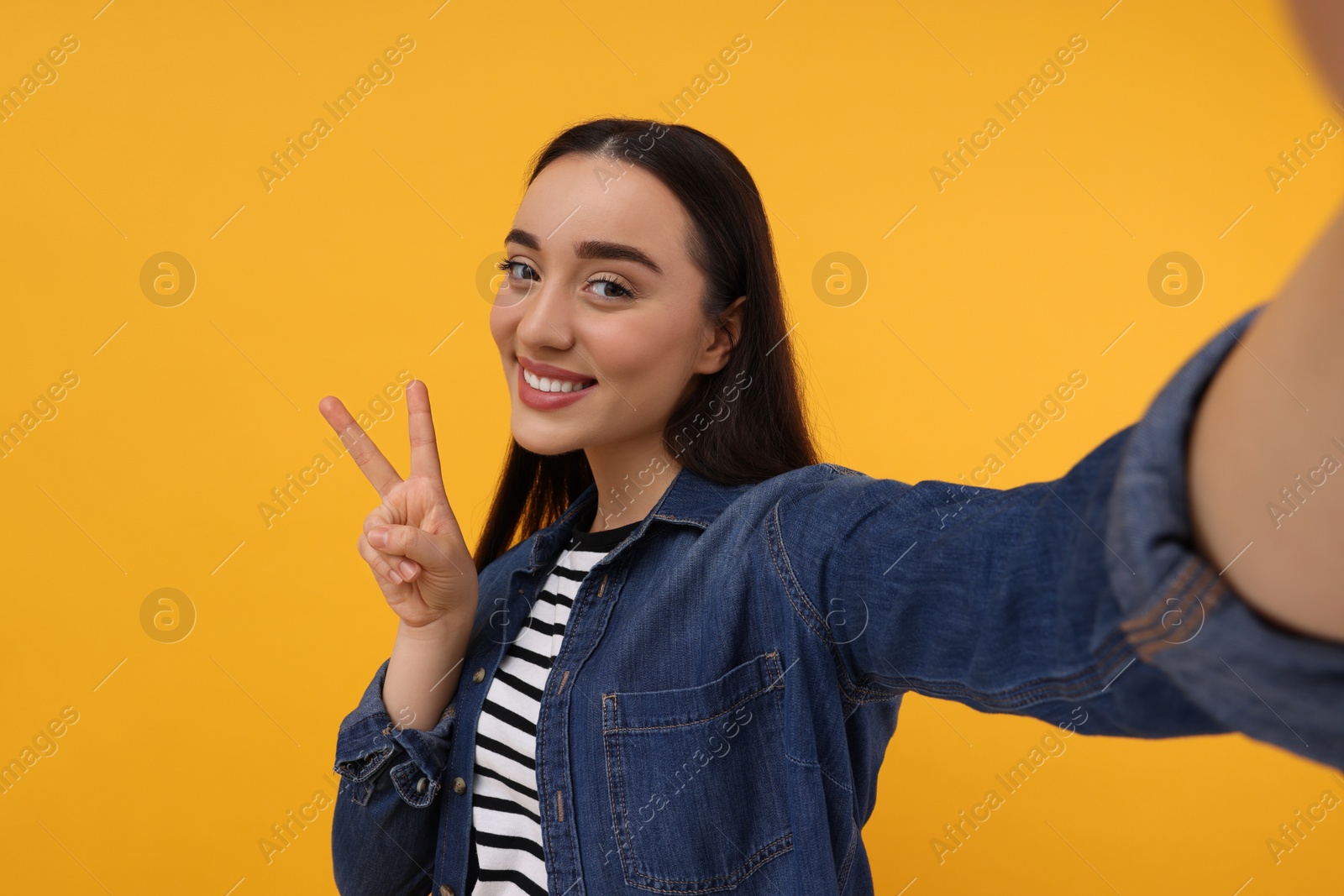Photo of Smiling young woman taking selfie and showing peace sign on yellow background