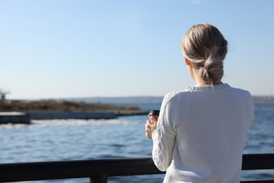 Lonely woman with cup of drink near river on sunny day, back view
