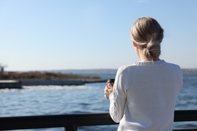 Photo of Lonely woman with cup of drink near river on sunny day, back view