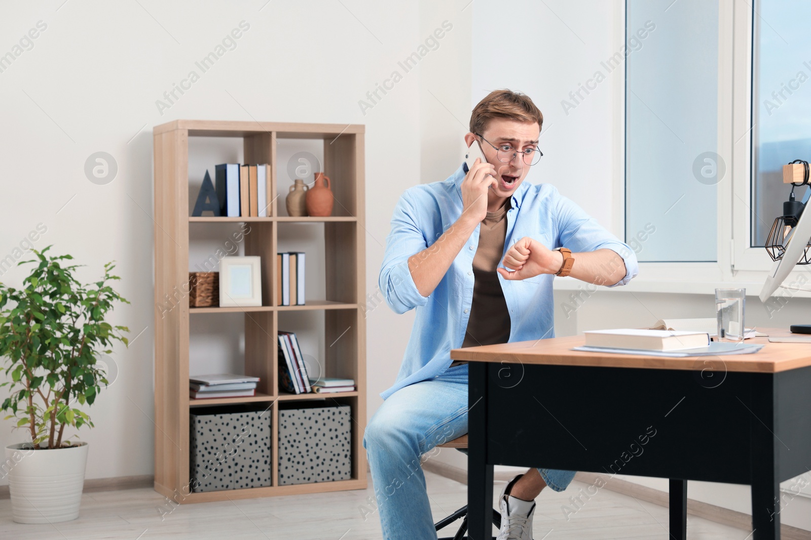 Photo of Emotional young man checking time while talking on phone in office. Being late