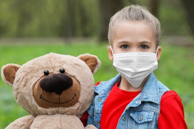 Photo of Little girl with protective mask and teddy bear outdoors
