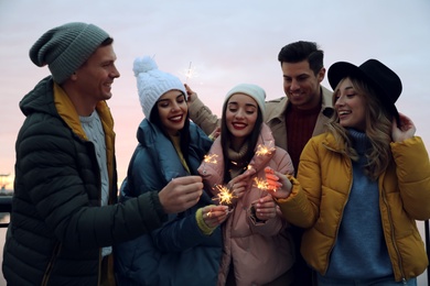 Photo of Group of people in warm clothes holding burning sparklers outdoors