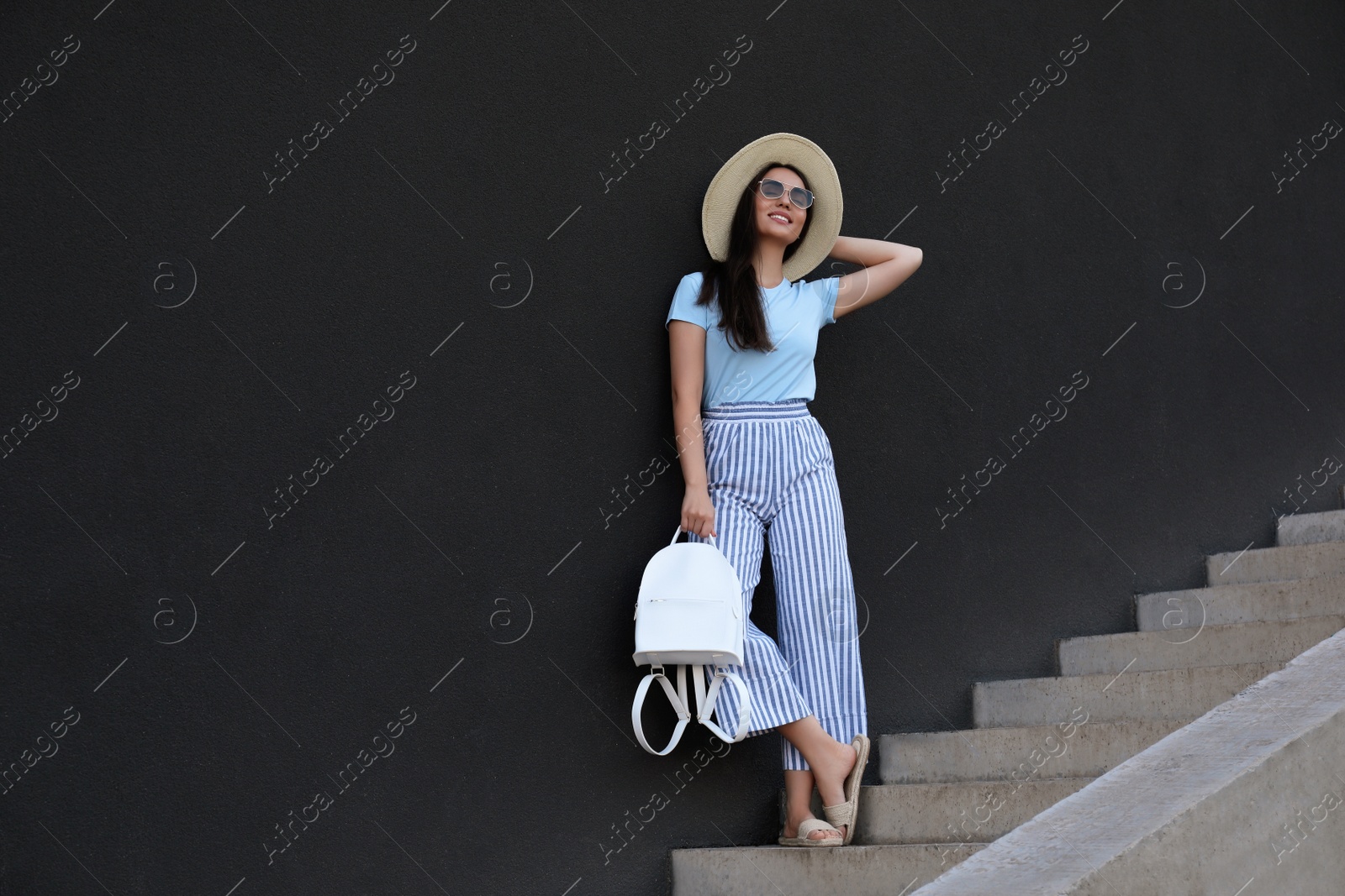 Photo of Beautiful young woman with stylish backpack and hat on stairs leaning to black wall