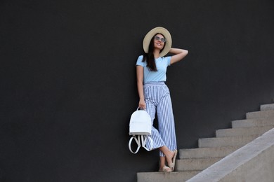 Photo of Beautiful young woman with stylish backpack and hat on stairs leaning to black wall