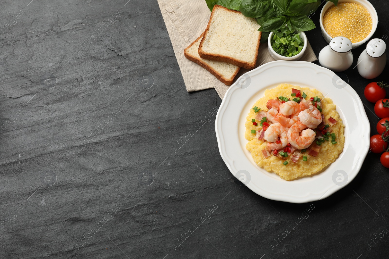 Photo of Plate with fresh tasty shrimps, bacon, grits, green onion and pepper on black textured table, flat lay. Space for text