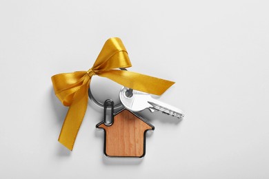 Key with trinket in shape of house and yellow bow on light grey background, top view. Housewarming party