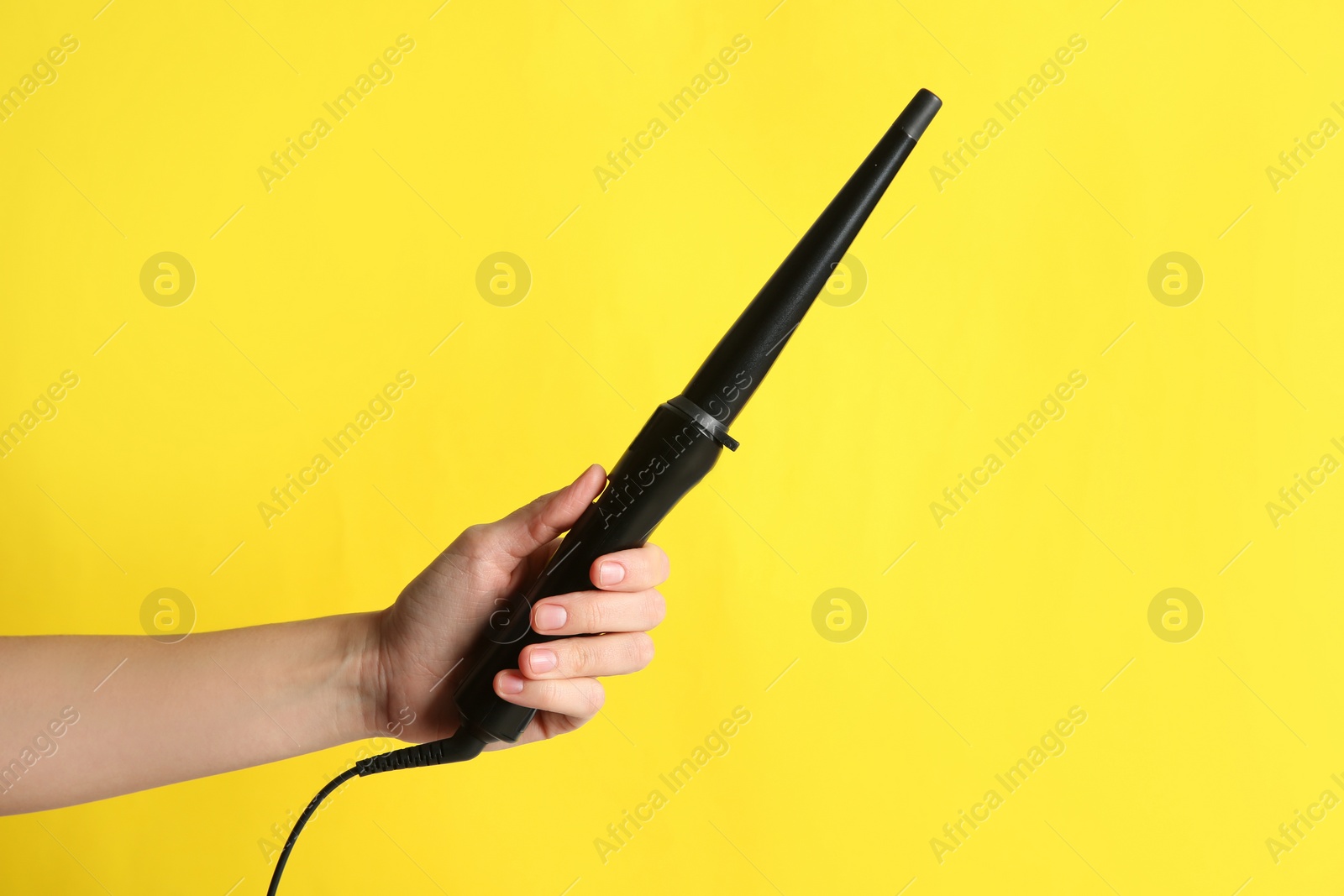 Photo of Woman holding clipless curling hair iron on yellow background, closeup