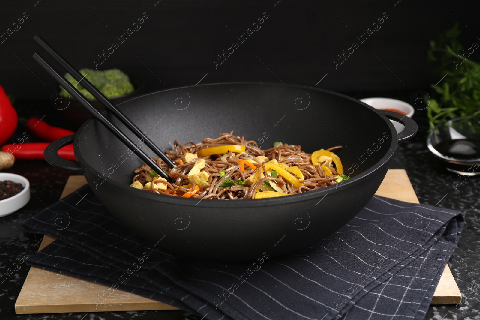 Photo of Stir-fry. Tasty noodles with vegetables, meat and chopsticks in wok on dark textured table