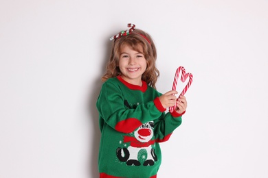 Cute little girl in Christmas sweater making heart shape with candy canes against white background