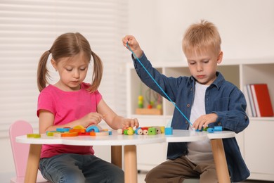 Little children playing with wooden pieces and string for threading activity at white table indoors. Developmental toys