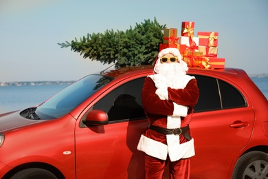 Photo of Authentic Santa Claus near car with presents and fir tree on roof outdoors