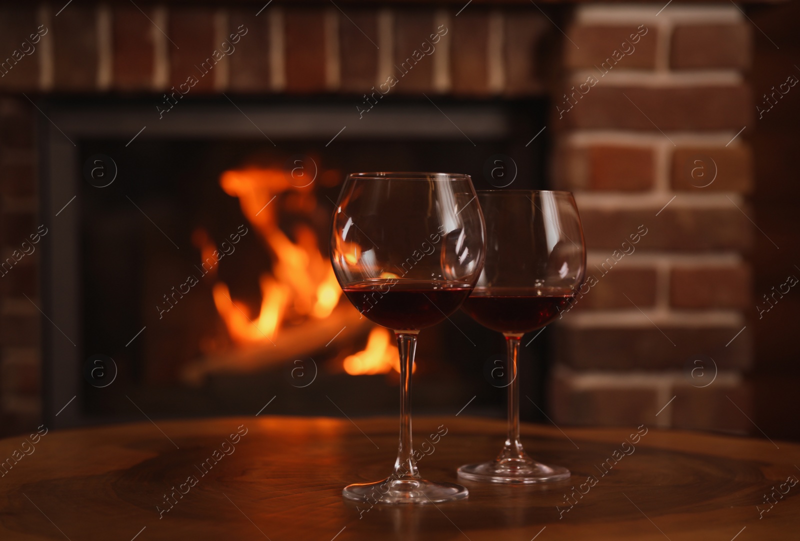 Photo of Glasses of red wine on wooden table near fireplace. Space for text