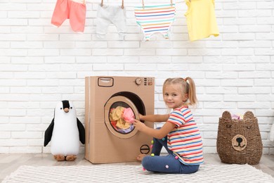Photo of Little girl putting laundry into toy cardboard washing machine indoors