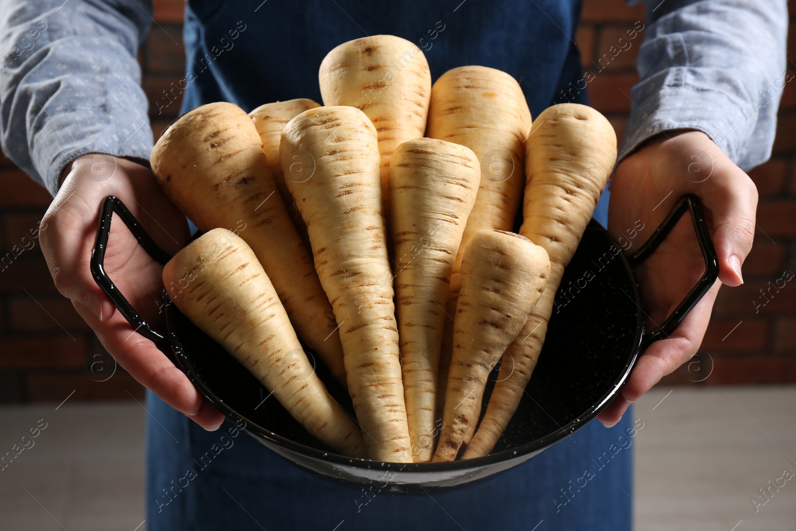 Photo of Woman holding bowl with delicious fresh ripe parsnips near red brick wall, closeup