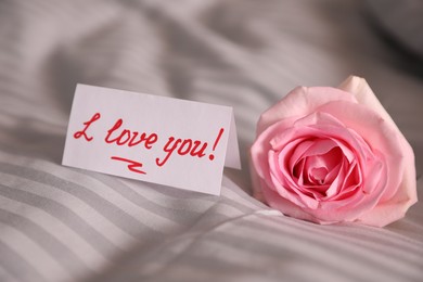 Photo of Note with handwritten text I Love You near pink rose on bed, closeup. Romantic message