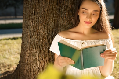 Beautiful young woman reading book near tree in park