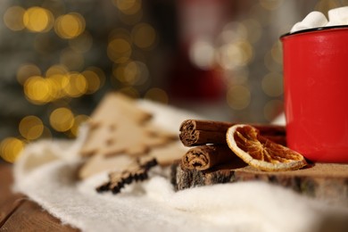 Cup of cocoa with marshmallows, cinnamon sticks, dry orange slices and Christmas decor on wooden table against festive lights, closeup. Space for text