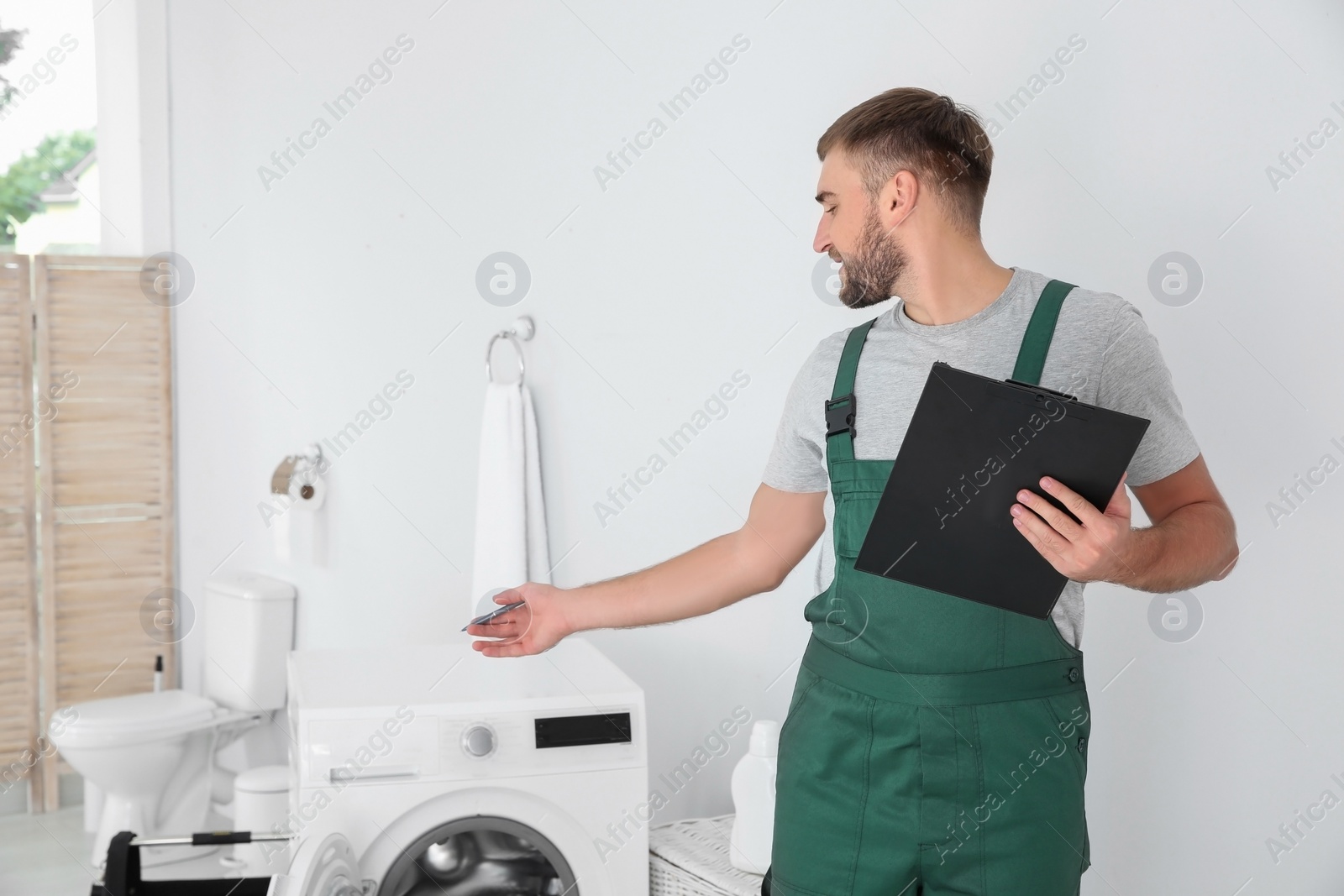 Photo of Young plumber with clipboard near washing machine in bathroom