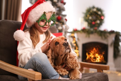 Photo of Cute little girl in Santa hat and Christmas glasses with English Cocker Spaniel at home