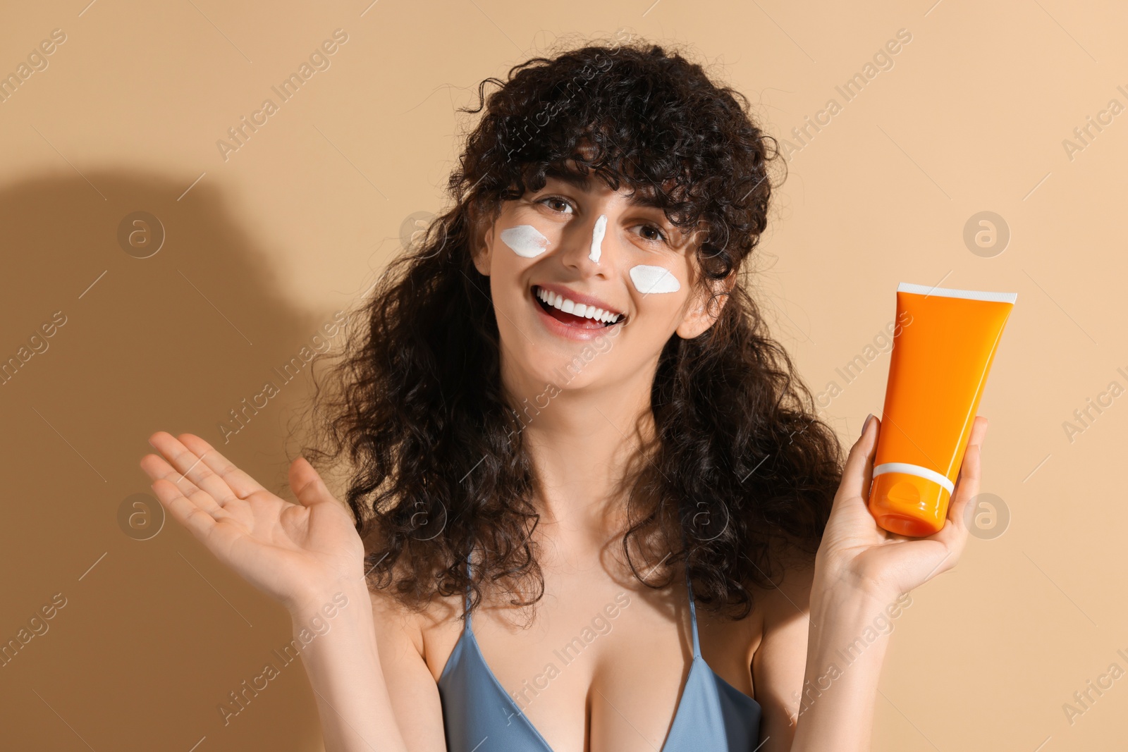 Photo of Beautiful young woman with sun protection cream on her face holding tube of sunscreen against beige background