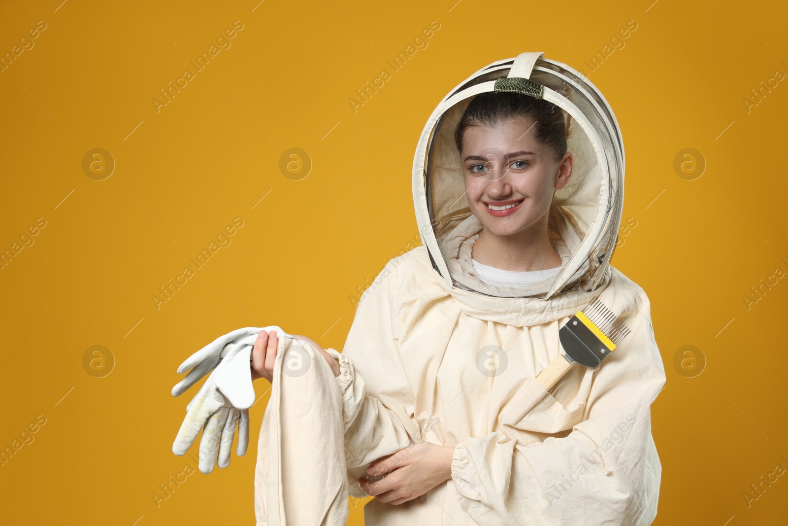 Photo of Beekeeper in uniform with gloves and uncapping fork on yellow background