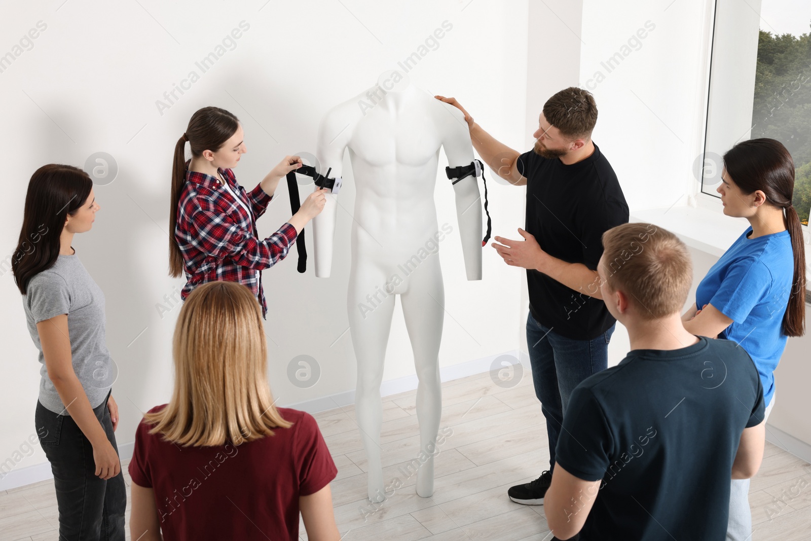 Photo of Group of people learning how to apply medical tourniquet on mannequin indoors