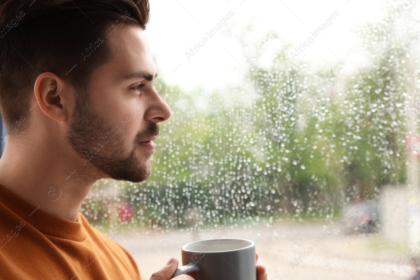 Photo of Thoughtful handsome man with cup of coffee near window indoors on rainy day