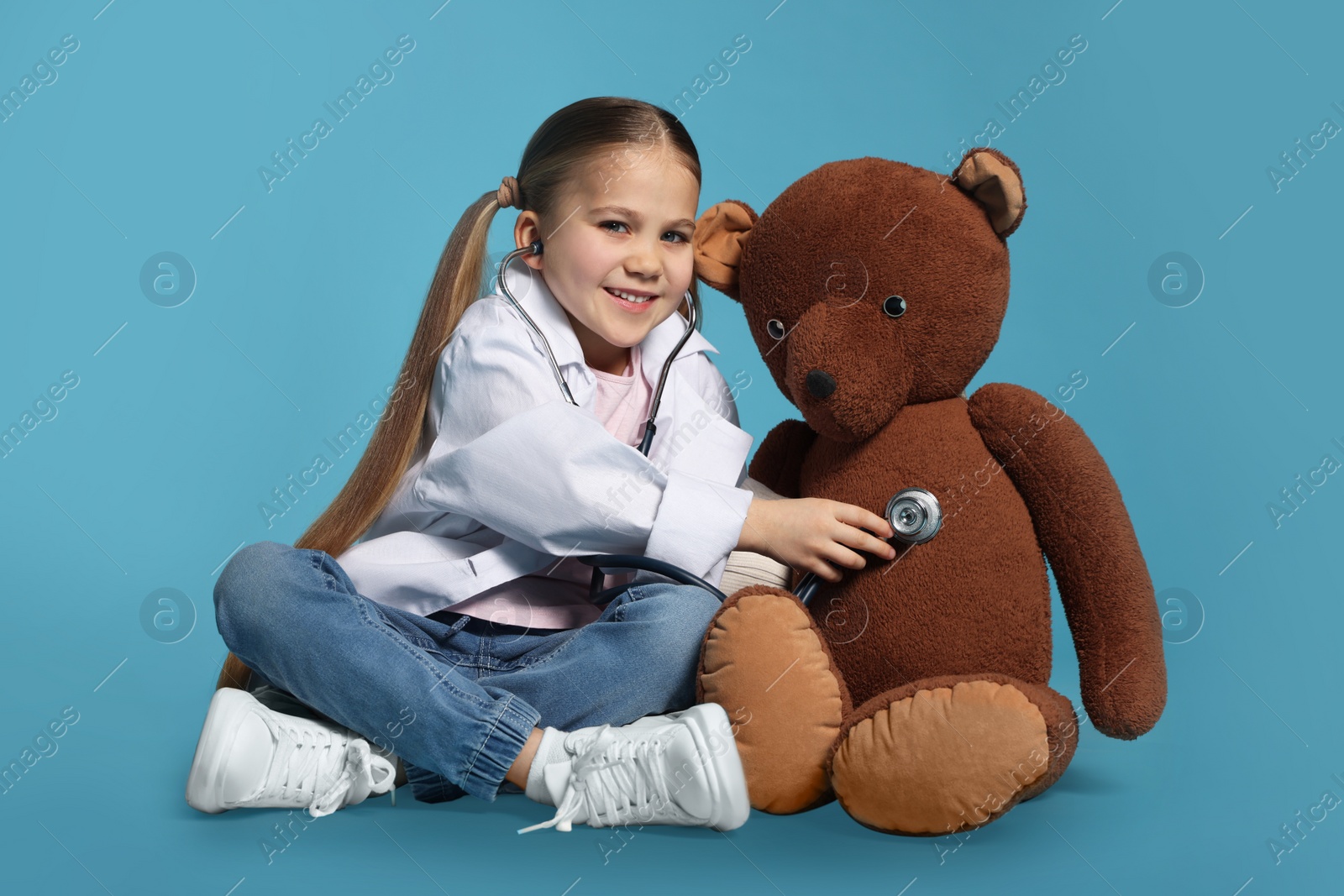 Photo of Little girl in medical uniform examining toy bear with stethoscope on light blue background