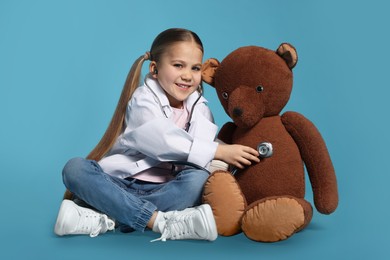 Little girl in medical uniform examining toy bear with stethoscope on light blue background