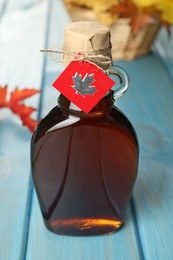 Glass bottle of tasty maple syrup and dry leaves on light blue wooden table, closeup