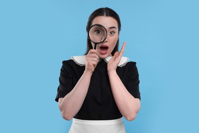 Photo of Emotional young woman in maid outfit looking through magnifier glass on light blue background