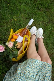 Photo of Woman sitting near yellow wicker bag with roses, wine, peaches and baguette on green grass outdoors, above view