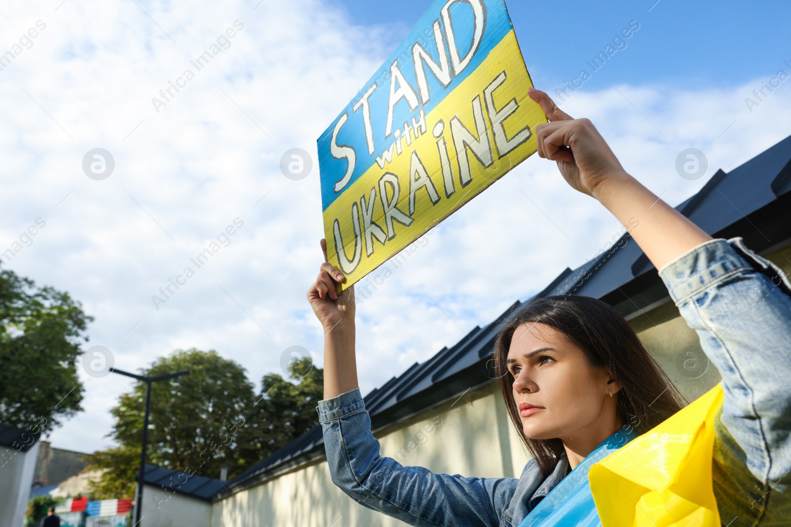 Photo of Sad woman holding poster in colors of national flag and words Stand with Ukraine outdoors. Space for text