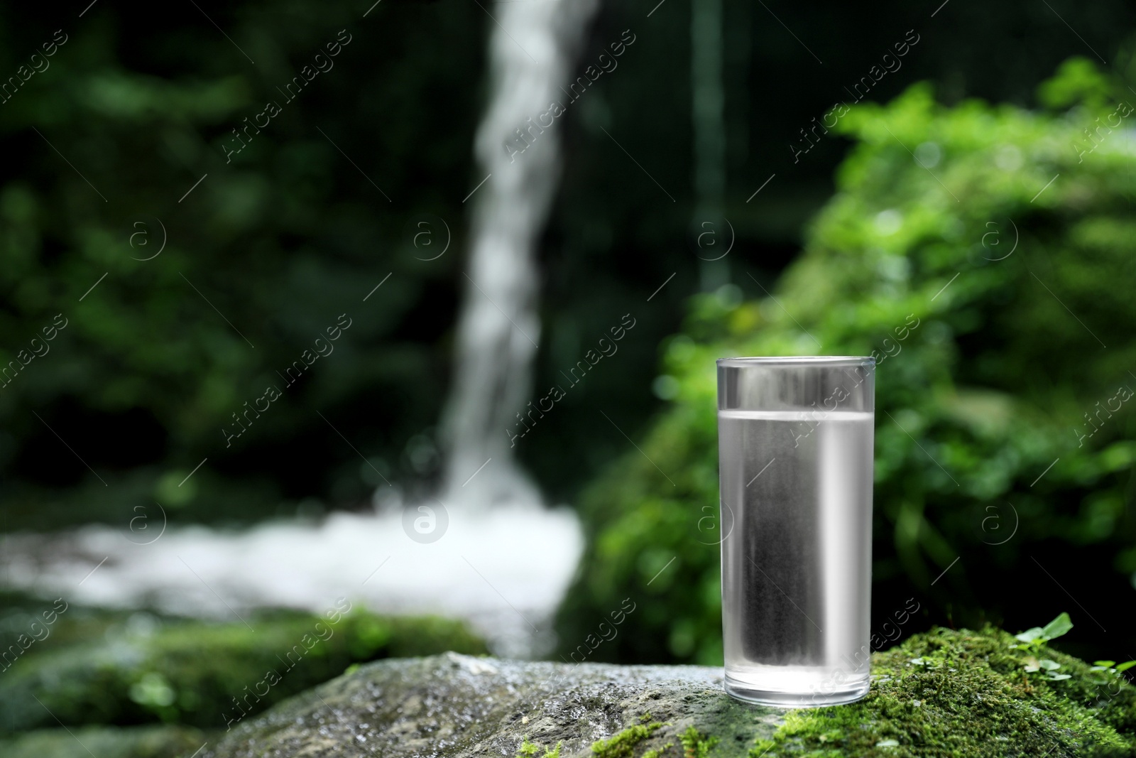 Photo of Glass of clean water near mountain waterfall outdoors. Space for text