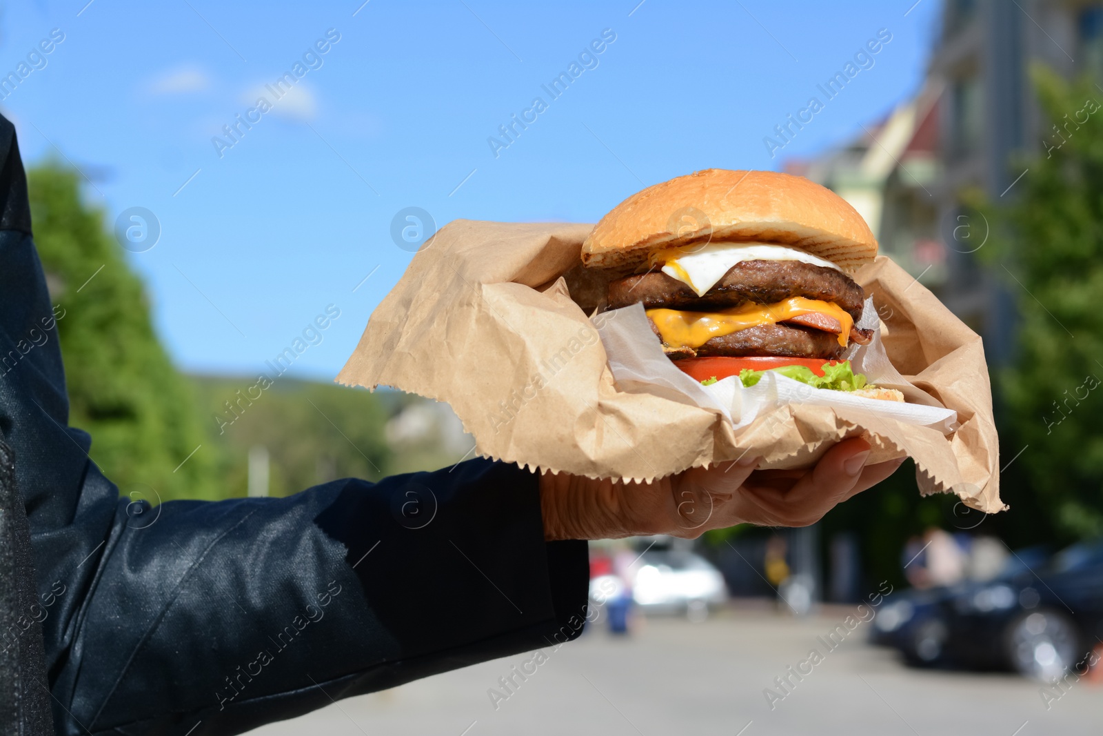 Photo of Woman holding delicious burger in paper wrap on city street, closeup